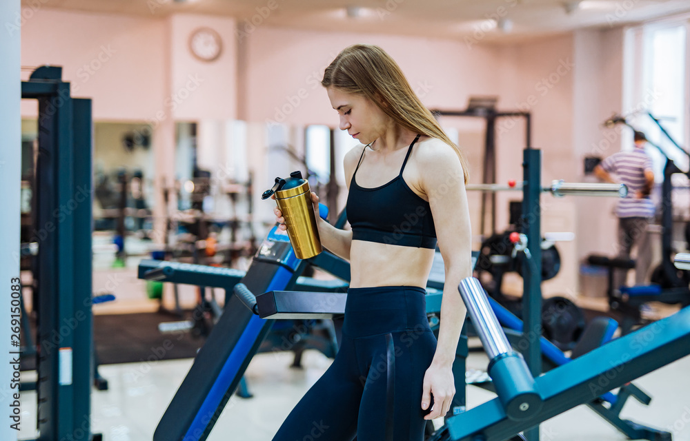 Beautiful female with muscular body is holding shaker and looking down in the gym. Slim sportswoman is trying to drink water after workout in the background of sport equipment indoors. Beautiful female with muscular body is holding shaker and looking down in the gym. Slim sportswoman is trying to drink water after workout in the background of sport equipment indoors.