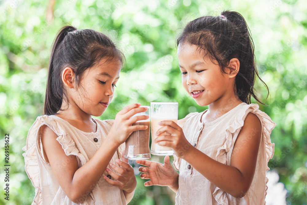 Two cute asian child girls drinking a milk from glass together on green nature background
