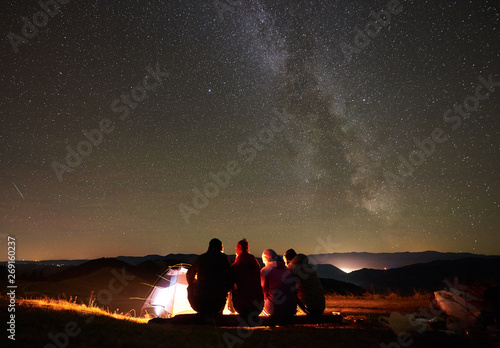 Night summer camping in mountains. Back view group of four hikers sitting on a bench made of logs together around bonfire near glowing tourist tent under night starry sky full of stars and Milky way.