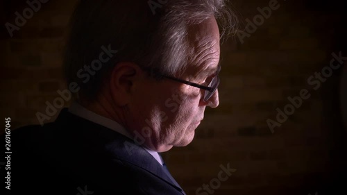 Close-up shot of silhouette of senior man in costume watches dramatically standing in complete darkness.