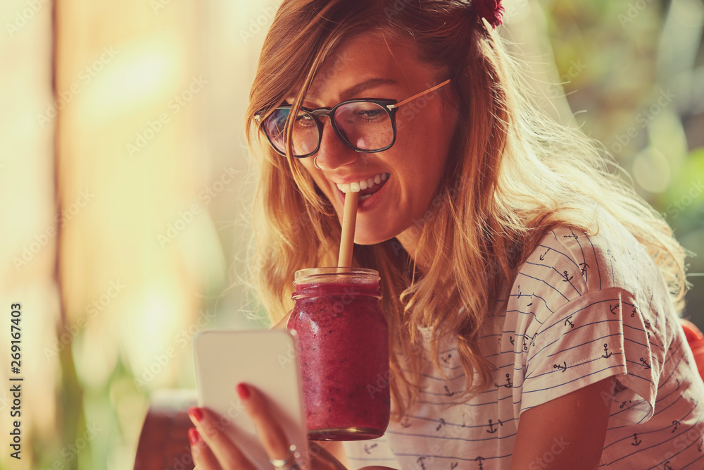 Beautiful woman using cellphone while drinking smoothie outdoors. - obrazy, fototapety, plakaty 