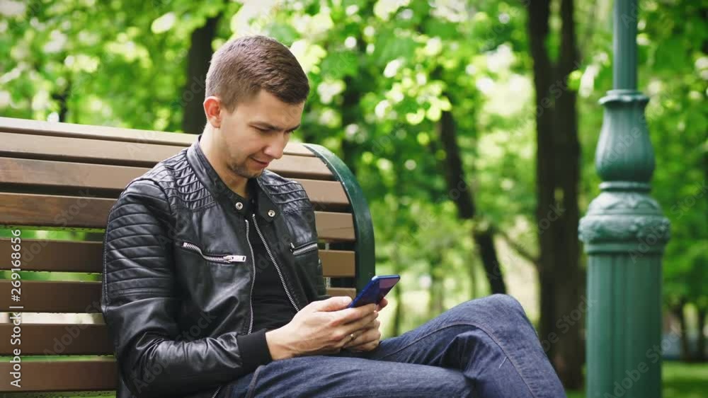 Attractive guy using a smartphone while sitting on a bench in the park Attractive guy using a smartphone while sitting on a bench in the park