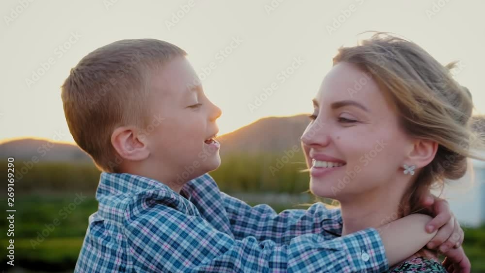 Engaging blonde child kissing with love his young smiling mother at countryside village, portrait of joyful family moment at green field on the warm sunset background, boy dressed in cage shirt Engaging blonde child kissing with love his young smiling mother at countryside village, portrait of joyful family moment at green field on the warm sunset background, boy dressed in cage shirt