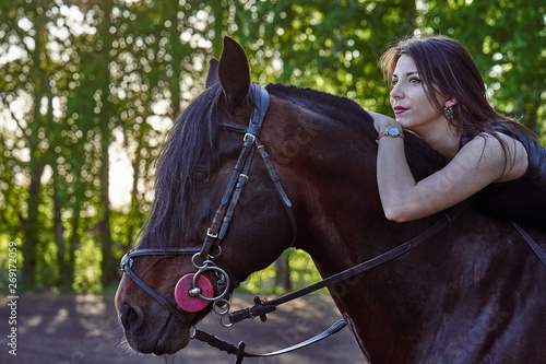 Young slim woman brunette in a black dress sitting on dark brown horse. Sunny summer evening. Close-up.