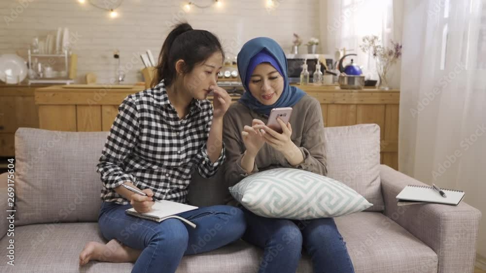 happy relax multi female friendship at home. malay woman talking telling news on smart phone sharing with korean girl who hard working doing homework on couch. two asian ladies discuss on cellphone happy relax multi female friendship at home. malay woman talking telling news on smart phone sharing with korean girl who hard working doing homework on couch. two asian ladies discuss on cellphone