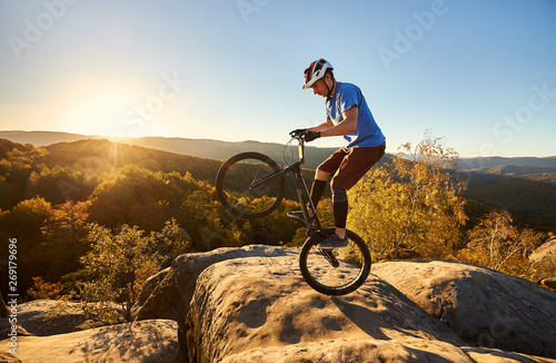 Young cyclist standing on back wheel on trial bicycle. Sportsman rider making acrobatic stunt on the edge of big boulder on the top of mountain at sunset. Concept of extreme sport active lifestyle