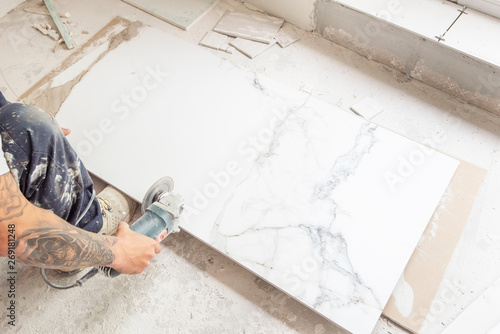 Tiler cutting a floor tile with a portable angle grinder. Craftsman hands using electric saw on marble ceramic tiles. Bathroom renovation concept