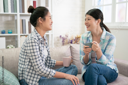 Happy asian sisters rest on sofa in living room talking planning future life holds cups drinking tea. two young girls best friends in shirt and jeans chatting laughing with hot coffee cup in hands.