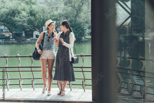 Photography Young joyful girls friends leaning on railing of wooden gazebo standing by pond in park with different trees