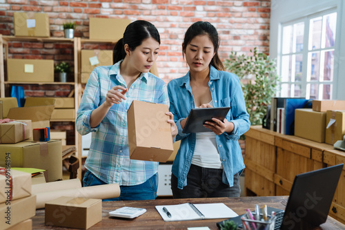Young women teamwork partners packing order into box together using touchpad to do delivery to client. two asian female colleagues with tablet checking goods number and prepared cardboards in office. © PR Image Factory