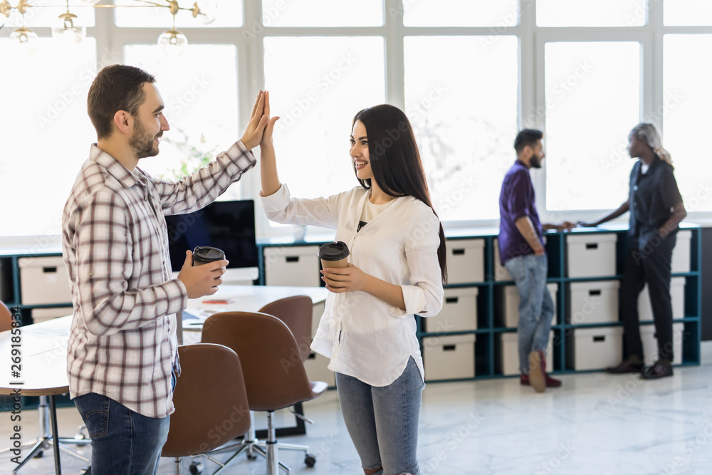 Give me high-five. Two coworkers giving high-five and holding coffee cups with smile while sitting on the couch at office with their beautiful coworker Give me high-five. Two coworkers giving high-five and holding coffee cups with smile while sitting on the couch at office with their beautiful coworker
