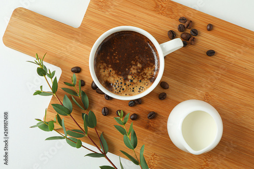 Cutting board with cup of fresh coffee, milk, coffee beans and plant branch on white background, top view