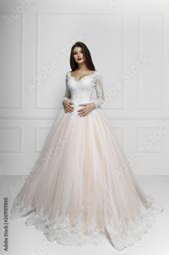 Frontal view of a fashion model in long elegant wedding dress, isolated on white background, near on white neoclassical wall, with copy space.