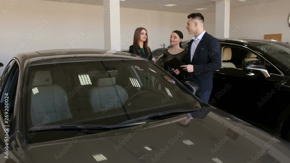 A successful car salesman tells two girls about the advantages of the car in a modern showroom. Two sisters choose a new car to buy. A successful car salesman tells two girls about the advantages of the car in a modern showroom. Two sisters choose a new car to buy.