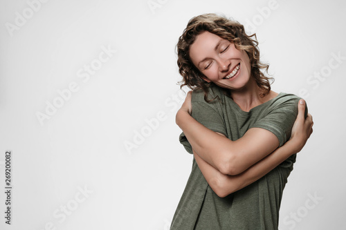 Canvastavla Young curly woman hugging herself, looks happy, expresses natural positive emoti
