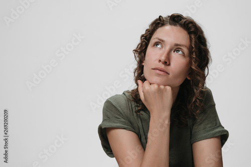 Young curly caucasian woman keeps hand under chin, being deep in thoughts