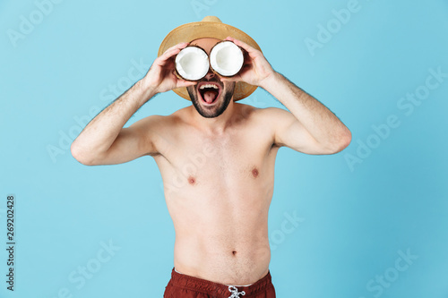 Photo of masculine shirtless tourist man wearing straw hat smiling while holding two coconut parts
