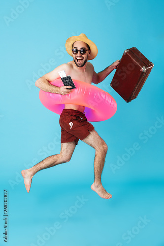 Photo of cheery shirtless tourist man wearing rubber ring holding travel tickets with passport and carrying luggage