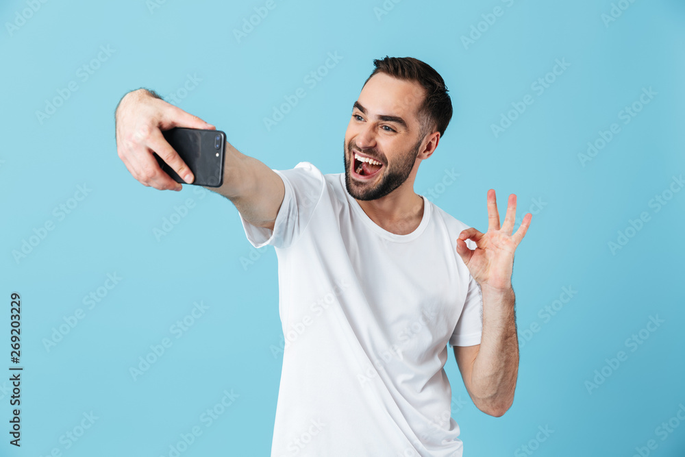Photo of positive brunette man wearing basic t-shirt laughing and taking selfie on smartphone Photo of positive brunette man wearing basic t-shirt laughing and taking selfie on smartphone