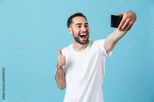 Photo of young brunette man wearing basic t-shirt laughing and taking selfie on smartphone