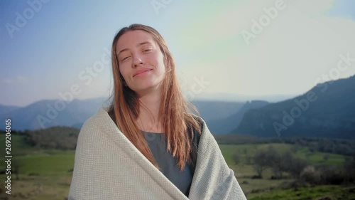 Nice Caucasian female smiles to camera and gently touching sunny long hair at green hill, eco friendly village girl standing with magic foggy mountains and field on the background and wrapping cosely photo