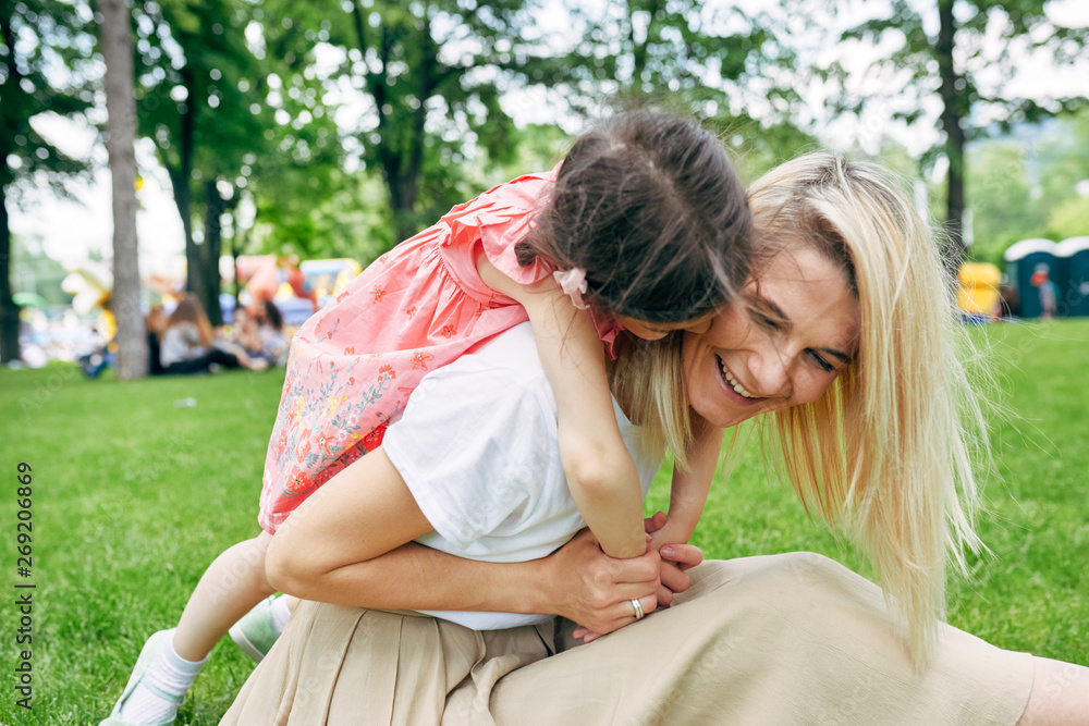 Cheerful child embracing mother in the park. Pretty daughter piggybacks her young attractive mother sitting on green grass outside. Love emotion. Happy Mothers Day. Motherhood and childhood concept Cheerful child embracing mother in the park. Pretty daughter piggybacks her young attractive mother sitting on green grass outside. Love emotion. Happy Mothers Day. Motherhood and childhood concept