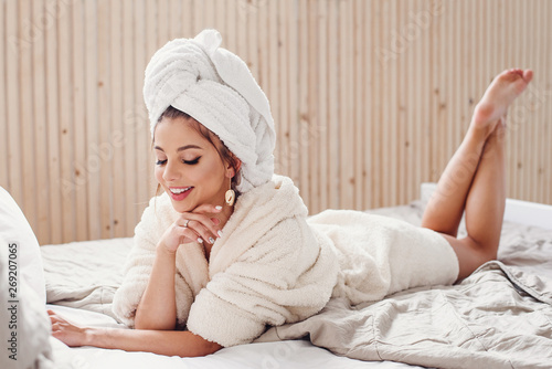 Pretty happy young woman on white bathrobe and with towel on her head lies on the bed at cozy home. Beautiful girl having relax on the bed in morning.
