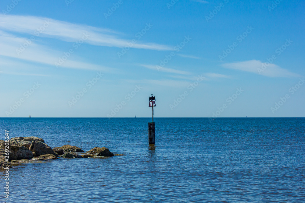 Fototapeta premium A view of a weathercock in the end of a stony groyne (breakwater) with a cormorant spreading his wing and crystal blue water under a majestic blue sky and some white clouds