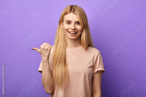 Optimistic young charming girl with toothy smile points aside with thumb, shows space for advertising. close up portrait.hurry up