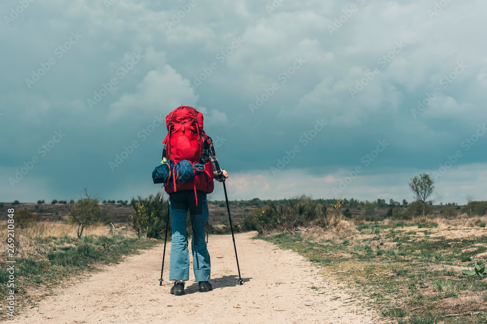 Backpacker going downhill on dirt road under cloudy sky.