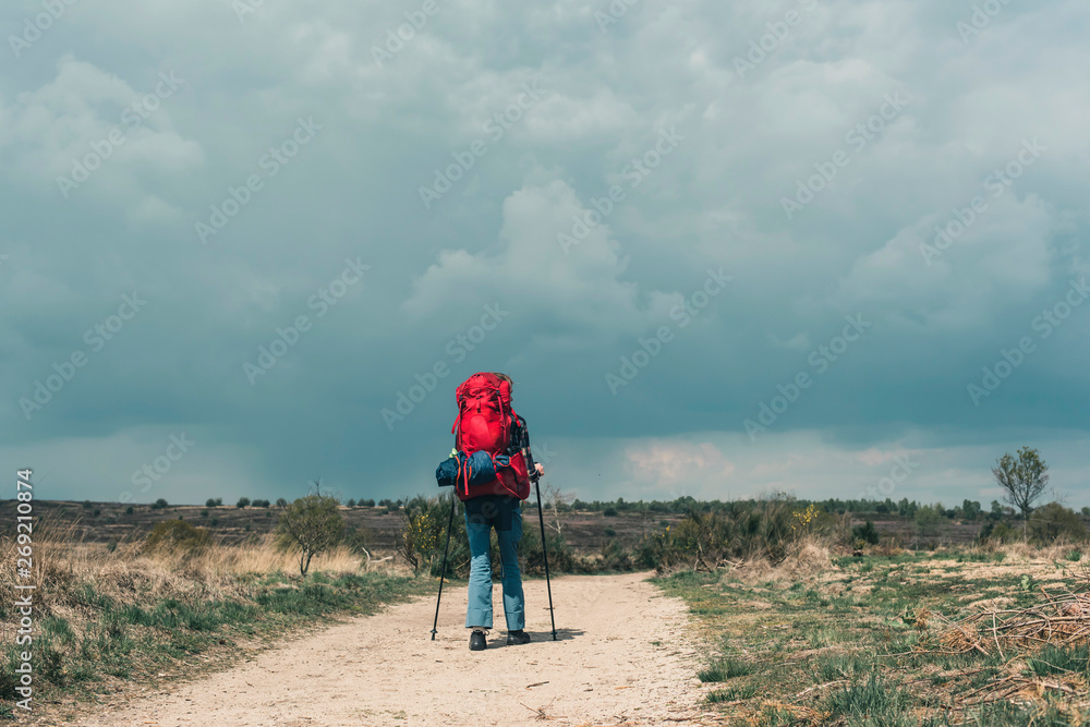 Backpacker going downhill on dirt road under cloudy sky.