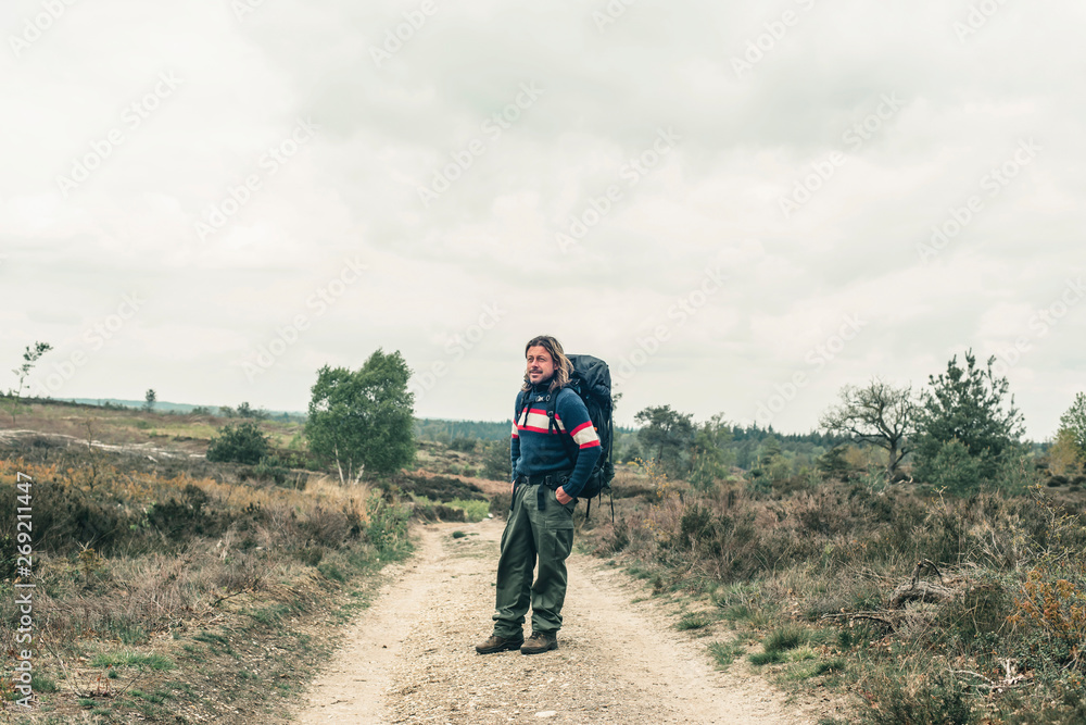 Man with long blonde hair and red backpack on dirt road in hilly landscape under cloudy sky.