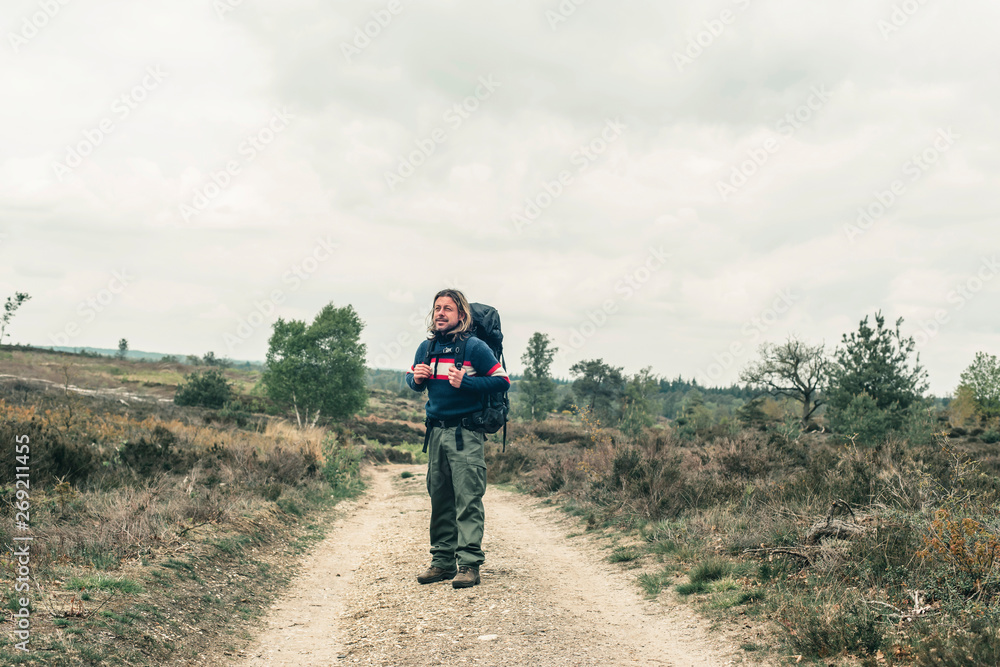 Man with long blonde hair and red backpack on dirt road in hilly landscape under cloudy sky.