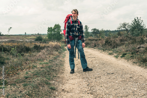 Young man with red backpack on dirt road in hilly landscape under cloudy sky.