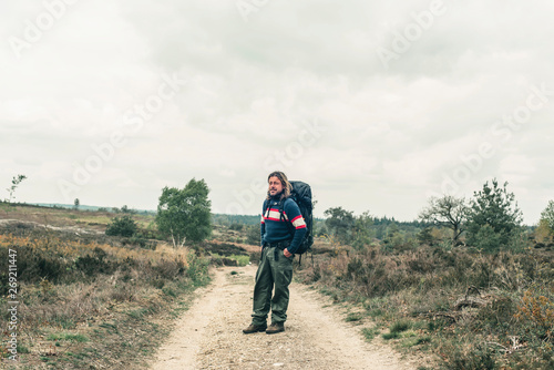 Man with long blonde hair and red backpack on dirt road in hilly landscape under cloudy sky.