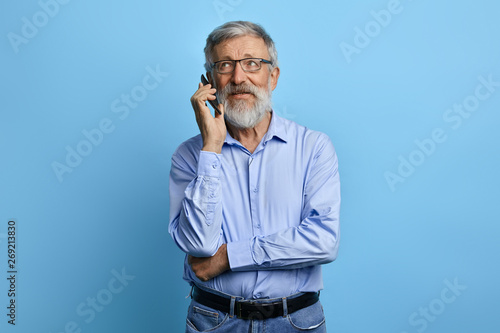 handsome senior man looking up, talking on mobile phone isolated against sky blue background. old businessman having chat, business conversation witj clients, parters. gadget concept. photo