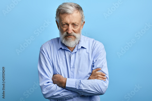 Handsome senior man in blue shirt with skeptic, nervous expression standing with crossed arms. close up portrait. isolated light blue background photo