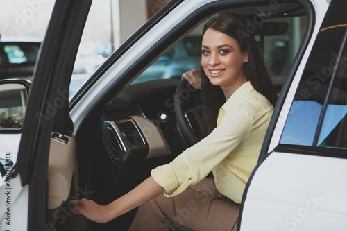 Excited young woman smiling to the camera, getting in a new automobile at the dealership. CHeerful beautiful woman sitting in a new car, choosing auto to buy at cars salon © Ihor