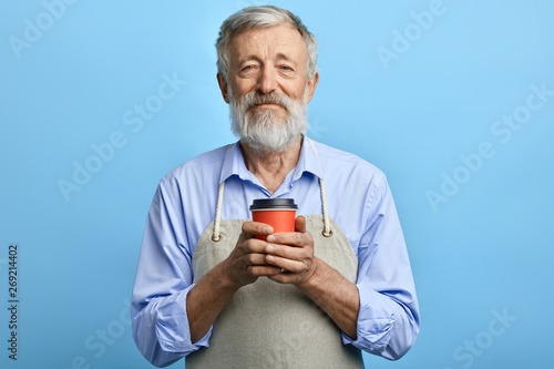 Friendly man in gray apron holding disposable cup of hot beverage. old waiter offers drink for client. business, service concept. would you like a cup of tea 