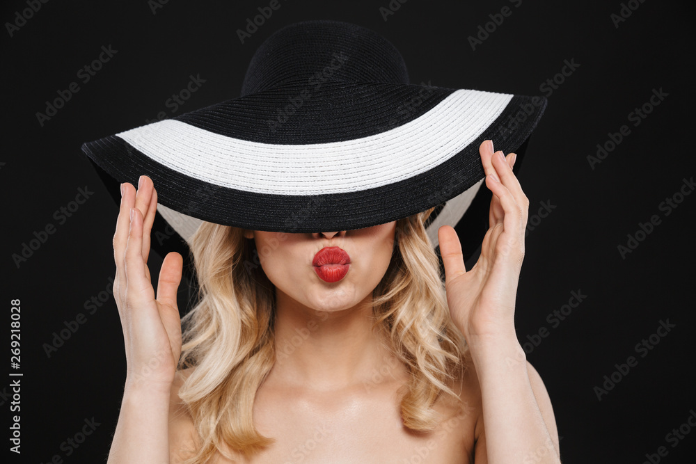 Young blonde woman with bright makeup red lips posing isolated over black wall background wearing hat blowing kisses. Young blonde woman with bright makeup red lips posing isolated over black wall background wearing hat blowing kisses.
