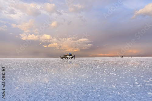 The tour group and car are in Salar de Uyuni during sunset. Beautiful clouds in the sunset light. In the foreground salt outgrowths in shallow water. Bolivia, South America