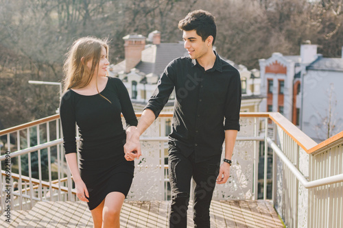 Beautiful young multiracial couple, a couple of students walk on the stairs in Kiev in the city center. Handsome Turkish brunette guy hugging, holding a hand Caucasian girl in black clothes