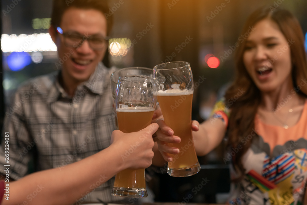 Group of asian friends holding pint glass of beer toasting for drink on Friday night in relaxing after work TGIF concept. Attractive young asian couple join happy hour night party at bar restaurant.