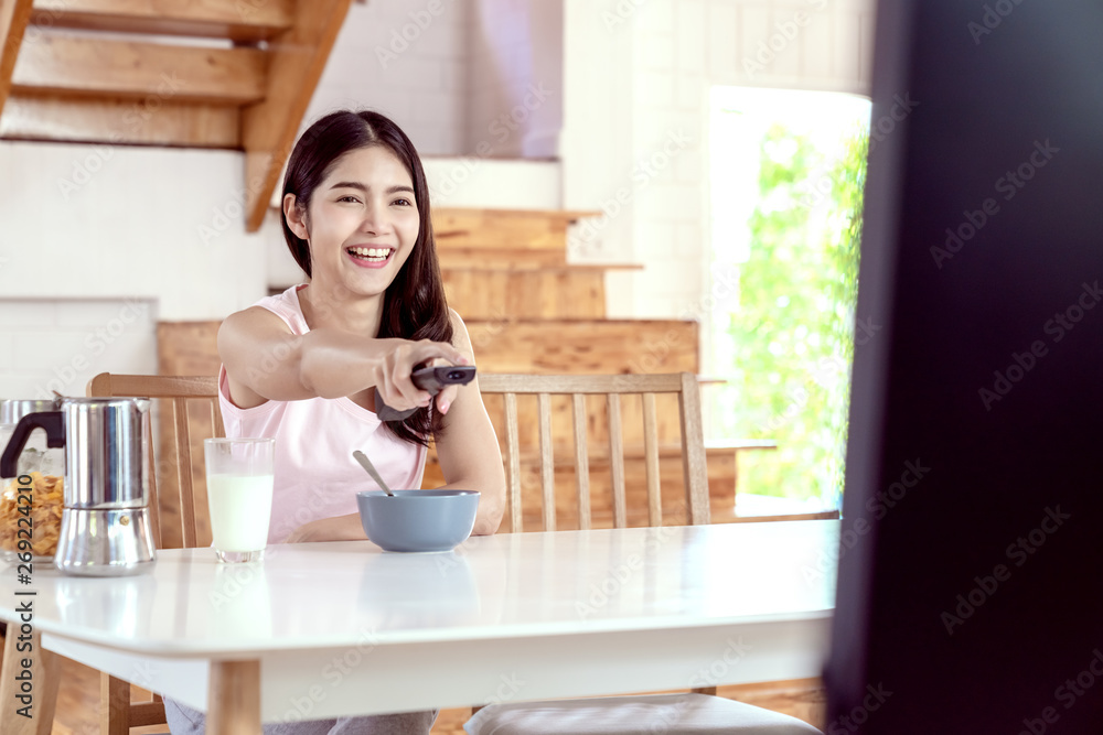 Candid of young happy attractive asian girl enjoy weekend activity by watching TV program at home. Young asian female student sitting on desk table holding remote on hand and pointing toward to TV. Candid of young happy attractive asian girl enjoy weekend activity by watching TV program at home. Young asian female student sitting on desk table holding remote on hand and pointing toward to TV.