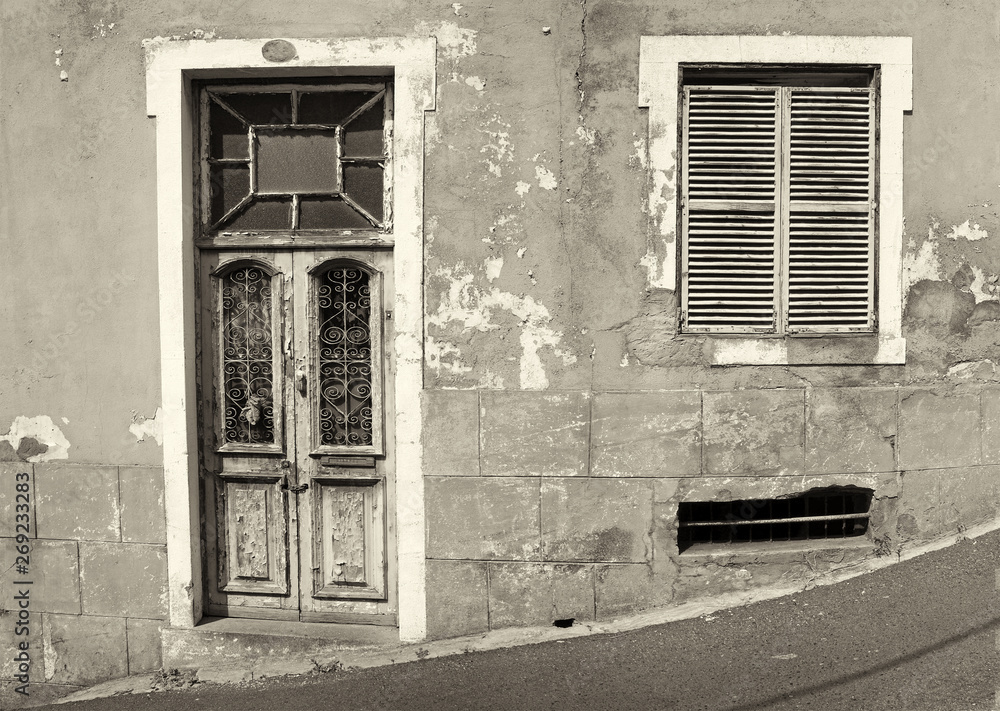 the front of an old abandoned house with shuttered windows and locked wooden door with flaking peeling paint on a sloping street monochrome image