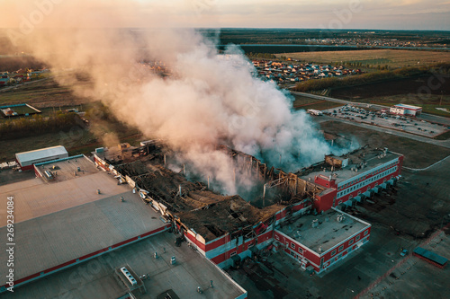 Aerial view of demolished burning industrial building by fire, huge smoke from broken roof, walls and piles of garbage