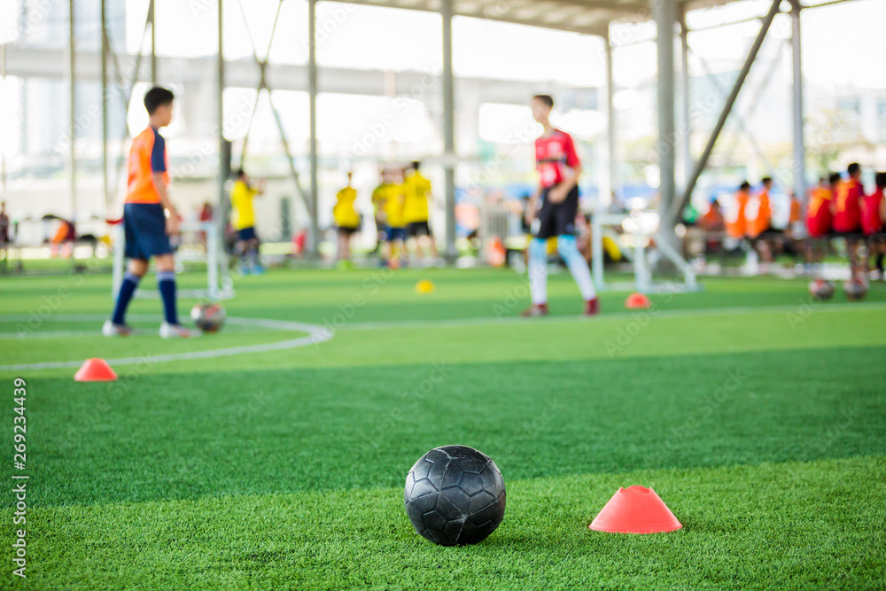 Fototapeta premium black football on green artificial turf with marker cone blurry soccer team training