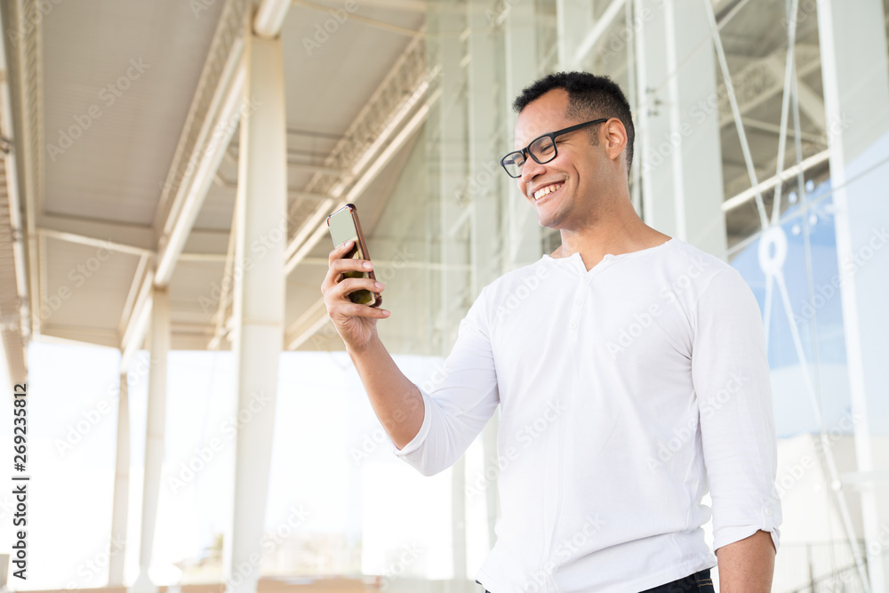 Bottom view of smiling young mixed-race man in spectacles and white T-shirt standing at office building, holding phone in hands, looking aside. Lifestyle concept Bottom view of smiling young mixed-race man in spectacles and white T-shirt standing at office building, holding phone in hands, looking aside. Lifestyle concept