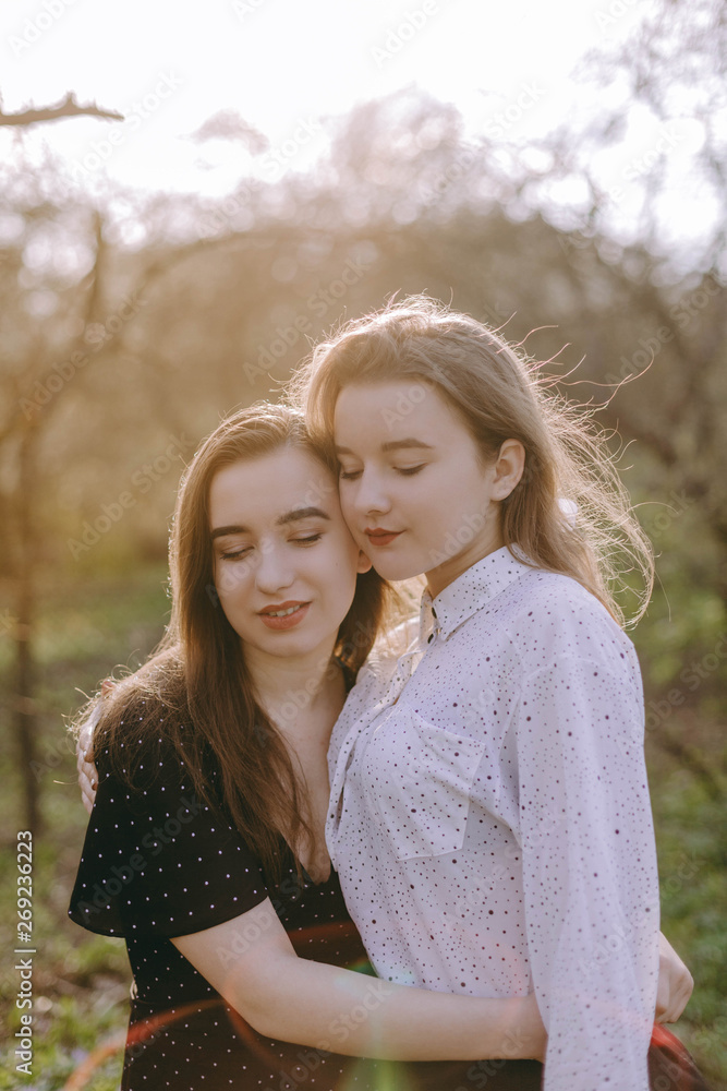 Fototapeta premium Portrait of two beautiful young sisters in the green spring garden, hugging in the field in the sunshine. Having fun together, positive emotions, bright colors. Copy space. Happy girlfriends at sunset