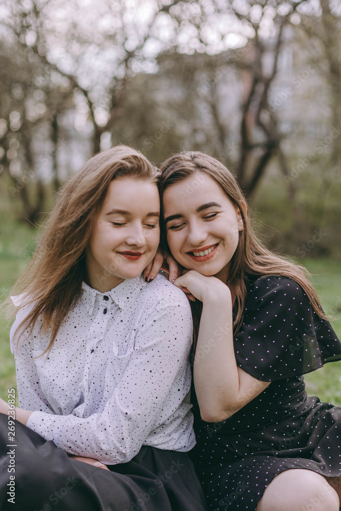 Fototapeta premium Portrait of two beautiful young sisters sitting in the green spring park, hugging in the field. Having fun together, positive emotions, bright colors. Copy space. Happy girlfriends at sunset